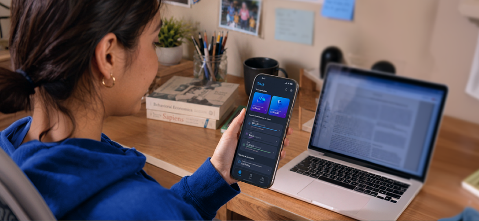 Uma estudante olhando a seção Track do app no celular, ao lado de um laptop.